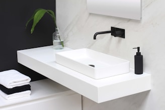 A minimalist bathroom sink setup featuring a rectangular white basin on a floating white countertop. A modern black faucet protrudes from a marble wall, complemented by a black soap dispenser. A small green plant in a glass vase and neatly stacked white and black towels add subtle accents.