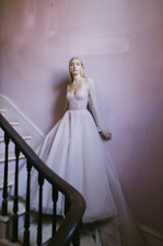 A woman in a sparkling, light-colored gown stands elegantly by a stairway. The flowing dress with sheer sleeves exudes sophistication. The stairwell features pale pink walls and a dark wooden banister, adding a touch of vintage charm.