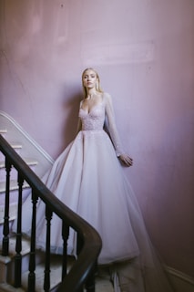 A woman in a sparkling, light-colored gown stands elegantly by a stairway. The flowing dress with sheer sleeves exudes sophistication. The stairwell features pale pink walls and a dark wooden banister, adding a touch of vintage charm.