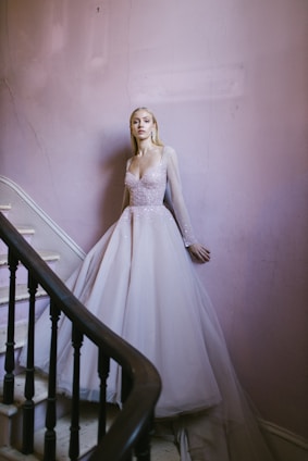 A woman in a sparkling, light-colored gown stands elegantly by a stairway. The flowing dress with sheer sleeves exudes sophistication. The stairwell features pale pink walls and a dark wooden banister, adding a touch of vintage charm.