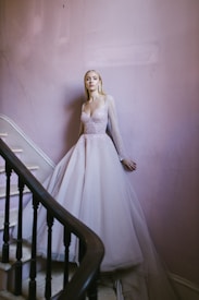 A woman in a sparkling, light-colored gown stands elegantly by a stairway. The flowing dress with sheer sleeves exudes sophistication. The stairwell features pale pink walls and a dark wooden banister, adding a touch of vintage charm.