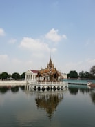 An ornate Thai-style pavilion is positioned on a serene body of water, surrounded by trees and additional buildings in the background. The pavilion reflects clearly in the calm water below.