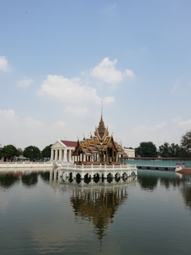 An ornate Thai-style pavilion is positioned on a serene body of water, surrounded by trees and additional buildings in the background. The pavilion reflects clearly in the calm water below.