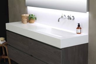 A modern bathroom sink setup features a sleek, white countertop with integrated basin and chrome wall-mounted faucet. On the left, a wicker basket and a small potted plant add natural elements. A brown soap dispenser sits on the right side of the sink. Below the countertop, wooden cabinets can be seen, and the background includes a large mirror reflecting a soft light.