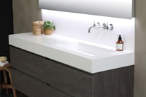 A modern bathroom sink setup features a sleek, white countertop with integrated basin and chrome wall-mounted faucet. On the left, a wicker basket and a small potted plant add natural elements. A brown soap dispenser sits on the right side of the sink. Below the countertop, wooden cabinets can be seen, and the background includes a large mirror reflecting a soft light.