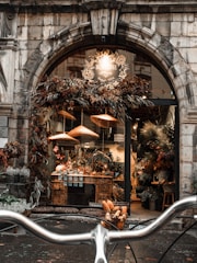 A rustic flower shop with a charming stone archway entrance, adorned with dried flowers and foliage. Inside, a variety of potted and fresh flowers are displayed on wooden tables. Soft, warm lights cast a cozy glow, enhancing the inviting ambience. In the foreground, bicycle handlebars are visible, suggesting a picturesque street setting.