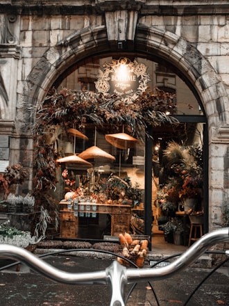 A rustic flower shop with a charming stone archway entrance, adorned with dried flowers and foliage. Inside, a variety of potted and fresh flowers are displayed on wooden tables. Soft, warm lights cast a cozy glow, enhancing the inviting ambience. In the foreground, bicycle handlebars are visible, suggesting a picturesque street setting.