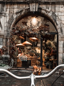 A rustic flower shop with a charming stone archway entrance, adorned with dried flowers and foliage. Inside, a variety of potted and fresh flowers are displayed on wooden tables. Soft, warm lights cast a cozy glow, enhancing the inviting ambience. In the foreground, bicycle handlebars are visible, suggesting a picturesque street setting.