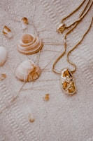Several seashells and a gold necklace are laid out on a textured beige fabric. The shells have a natural, earthy tone with various shades of brown and white. The necklace features a shiny, golden pendant with intricate details.