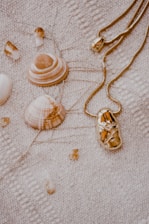 Several seashells and a gold necklace are laid out on a textured beige fabric. The shells have a natural, earthy tone with various shades of brown and white. The necklace features a shiny, golden pendant with intricate details.