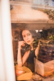 A person is sitting indoors at a round table, gazing thoughtfully through a window. The view is partially obscured by reflections. A closed book rests on the table, adding to the contemplative scene.