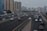 Multiple vehicles are traveling on a multi-lane highway with several buildings in the background. Streetlights illuminate the road, and the sky appears to be overcast. There is a variety of vehicles including cars and SUVs, and street signs are visible along the sides of the road.