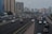 Multiple vehicles are traveling on a multi-lane highway with several buildings in the background. Streetlights illuminate the road, and the sky appears to be overcast. There is a variety of vehicles including cars and SUVs, and street signs are visible along the sides of the road.
