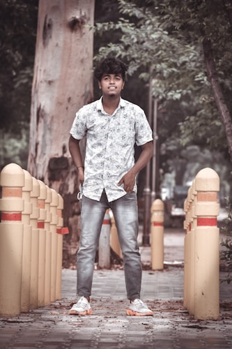 A person stands confidently on a pathway lined with yellow bollards. The person is wearing a floral shirt, jeans, and sneakers. The background includes trees and a large tree trunk, giving a natural, outdoor setting.