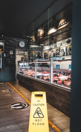 A delicatessen with a glass display containing various meats and cheeses. The floor is marked with social distancing stickers and a yellow caution sign indicating a wet floor. The decor features dark brick walls and overhead spotlighting. A large wall clock is visible near the entrance.