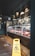 A delicatessen with a glass display containing various meats and cheeses. The floor is marked with social distancing stickers and a yellow caution sign indicating a wet floor. The decor features dark brick walls and overhead spotlighting. A large wall clock is visible near the entrance.