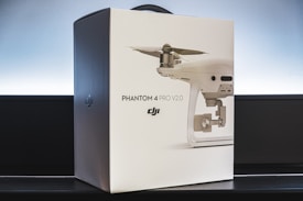 A box of the DJI Phantom 4 Pro V2.0 drone is placed on a flat surface. The box is mostly white with a printed image of the drone along with its branding and model information on the front. The background features a subtle gradient of light to dark, enhancing the sleek and modern presentation.