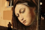 A detailed mural of a serene, contemplative face painted on a wall, featuring soft tones and realistic shading. The surrounding area includes a window and shadows that suggest evening light.