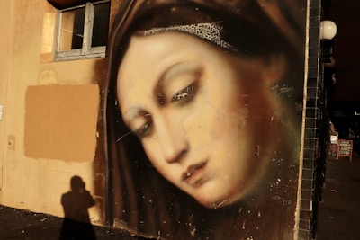 A detailed mural of a serene, contemplative face painted on a wall, featuring soft tones and realistic shading. The surrounding area includes a window and shadows that suggest evening light.