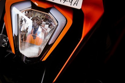 Close-up view of a motorcycle headlight with an angular design. The headlamp features a chrome reflective surface inside, and the surrounding structure is primarily orange with black accents. A portion of a license plate is visible above the headlight.