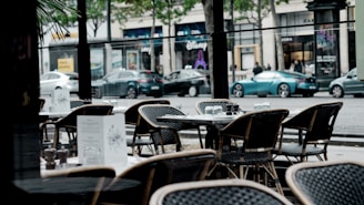 Outdoor seating area of a cafe with wicker chairs and tables set for dining, menus are placed on the tables. Behind, a city street with parked cars and shops in the background, surrounded by trees along the sidewalk.