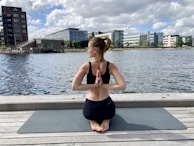 A person is practicing yoga on a wooden deck beside a body of water. They are sitting on a yoga mat in a cross-legged position with hands in a prayer pose behind their back. The backdrop includes modern buildings and cloudy sky.