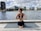 A person is practicing yoga on a wooden deck beside a body of water. They are sitting on a yoga mat in a cross-legged position with hands in a prayer pose behind their back. The backdrop includes modern buildings and cloudy sky.