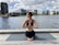 A person is practicing yoga on a wooden deck beside a body of water. They are sitting on a yoga mat in a cross-legged position with hands in a prayer pose behind their back. The backdrop includes modern buildings and cloudy sky.
