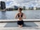 A person is practicing yoga on a wooden deck beside a body of water. They are sitting on a yoga mat in a cross-legged position with hands in a prayer pose behind their back. The backdrop includes modern buildings and cloudy sky.
