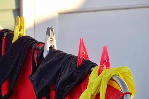 A collection of laundry items, including black and yellow fabric, are clipped on a metal railing using colorful clothespins. The clothespins are red and yellow, securing various textured clothes under bright lighting.