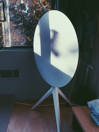 A modern white speaker with a tripod stand is positioned in a room next to a window. Natural light casts shadows on the speaker, creating an interesting play of light and shadow. Outside the window, tree branches with autumn leaves are visible, adding a touch of nature.