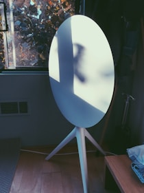 A modern white speaker with a tripod stand is positioned in a room next to a window. Natural light casts shadows on the speaker, creating an interesting play of light and shadow. Outside the window, tree branches with autumn leaves are visible, adding a touch of nature.