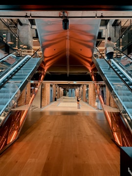 An architectural interior featuring two parallel escalators in a modern building with industrial design elements. The space includes exposed brick walls, a wooden floor, and soft lighting. A person is ascending one of the escalators while another stands at the far end of the room, giving a sense of depth and scale.