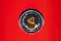 A close-up view of a metal bolt or screw embedded in a vibrant red surface. The metal is slightly rusted and set within a shiny, silver-colored circular frame.