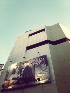 A tall building with a large advertisement for 'Dickies' featuring a man and woman on its facade. The billboard displays the brand logo and promotional text. The sky is clear with some exposed wiring attached to the structure.