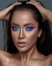 A person with striking makeup featuring vivid blue and purple eye shadow, wet hair, and a glistening complexion. The pose involves hands placed on the head, showcasing a confident and bold look.