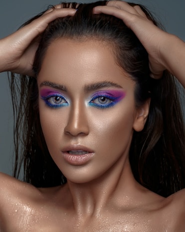 A person with striking makeup featuring vivid blue and purple eye shadow, wet hair, and a glistening complexion. The pose involves hands placed on the head, showcasing a confident and bold look.