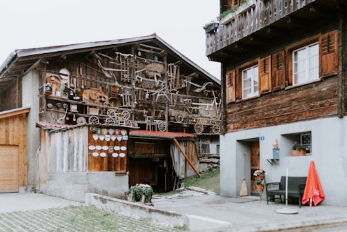 A rustic outdoor scene features two wooden buildings with a variety of old tools and wooden objects mounted on one of the facades. The buildings have wooden shutters and balconies with flower pots. A broom and a red umbrella adorn the entrance of one building, while a stone pathway and grass are visible in the foreground.