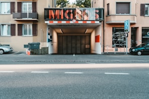A city street features an entrance to a parking garage labeled with large letters. Flanking the entrance are apartment buildings with balconies and windows. Vehicles are parked along the street, and there are signs indicating parking regulations. A trash bin and some traffic elements are visible in the surroundings.