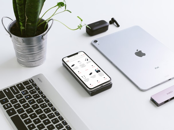 A clean workspace features a potted plant in a metal container, a smartphone displaying an online store, a laptop keyboard, a tablet, wireless earbuds, and a USB hub. The arrangement creates a modern, minimalist tech setup.