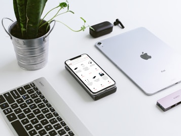 A clean workspace features a potted plant in a metal container, a smartphone displaying an online store, a laptop keyboard, a tablet, wireless earbuds, and a USB hub. The arrangement creates a modern, minimalist tech setup.