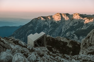 A small, modern cabin with a triangular roof is situated on a rocky mountain slope, surrounded by rugged peaks. The warm glow of the setting sun highlights the mountain ridges in the distance, casting a serene ambiance over the landscape.