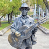 A detailed sculpture of a person playing an acoustic guitar is positioned in an outdoor setting. The figure is wearing a hat and a scarf, with a gentle smile on its face. The sculpture appears to be made of a metallic material, capturing intricate details of the figure's clothing and guitar. Surrounding the sculpture are trees and pathways, suggesting it is situated in a park or garden area.