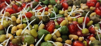 A close-up view of a mixture of sprouted seeds and pomegranate seeds. The image features different sizes and shapes of green, red, and beige sprouts intertwined, with white sprout shoots emerging from some of the green and beige seeds.