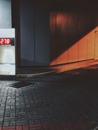 Dimly lit urban scene featuring a digital display board with a number 278, mounted against a textured wall with an orange-brown color leading to an entrance. The ground is paved with patterned tiles, and there's a gradient of light creating shadows and contrasts.
