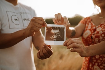 Two people are holding an ultrasound photo together, with one person wearing a white shirt and the other wearing a red floral dress. The scene is set outdoors, with a soft, warm lighting creating a serene atmosphere.