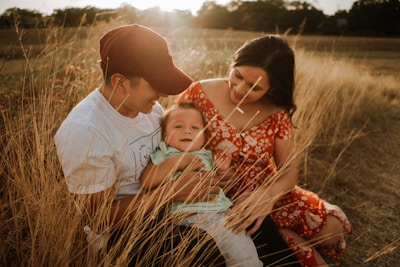 A family sits together in a field of tall grass during sunset, with a man in a red cap and white t-shirt holding a baby who is smiling. A woman in a floral red dress is sitting beside them, looking affectionately at the child.