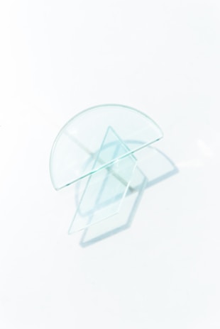 A minimalist composition featuring two transparent glass shapes, a semicircle and a triangle, arranged on a white background. The shapes cast soft shadows, creating an abstract and geometric design.