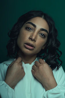 A person with curly dark hair stands against a dark green background, wearing a white garment. Their face is expressive, with visible freckles and a contemplative gaze. Both hands are raised to adjust the collar, creating a focused and emotionally intriguing moment.