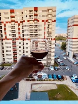 A hand holds a glass of red wine against a backdrop of a modern residential apartment complex. The buildings are characterized by white and red geometric patterns. Below, a parking lot with several cars and a small playground are visible.